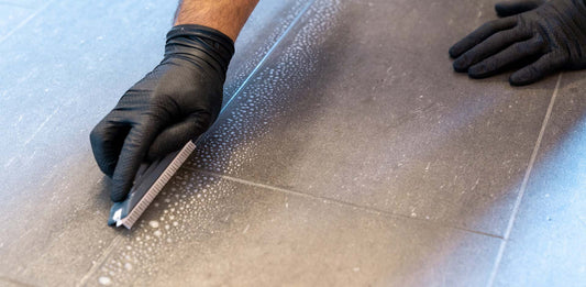 Gloved hand using a rubber float to tool grout into a tile joint, with grout residue splattered across the floor